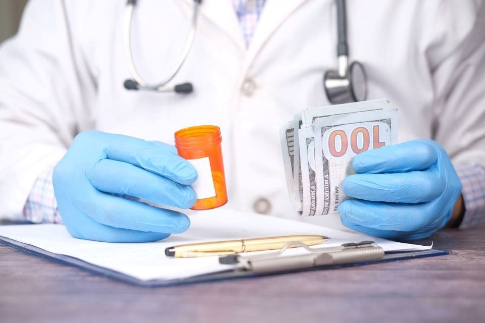 Doctor holding pills and money over a clipboard.