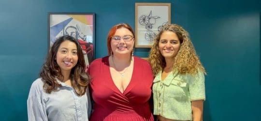 Three women standing side by side, smiling with art hung on a wall behind them.
