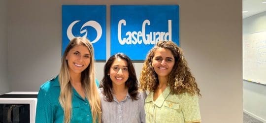 Three women standing side by side, smiling with CaseGuard art hung on a wall behind them.