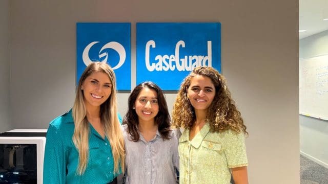 Three women standing side by side, smiling with CaseGuard art hung on a wall behind them.