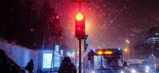 Crowded street with buses and cars. Snow is falling. There is a red traffic light glowing in the middle of the image.