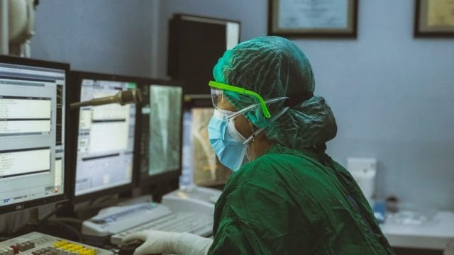 Doctor in green scrubs looking at multiple computer screens with sensitive medical information on them